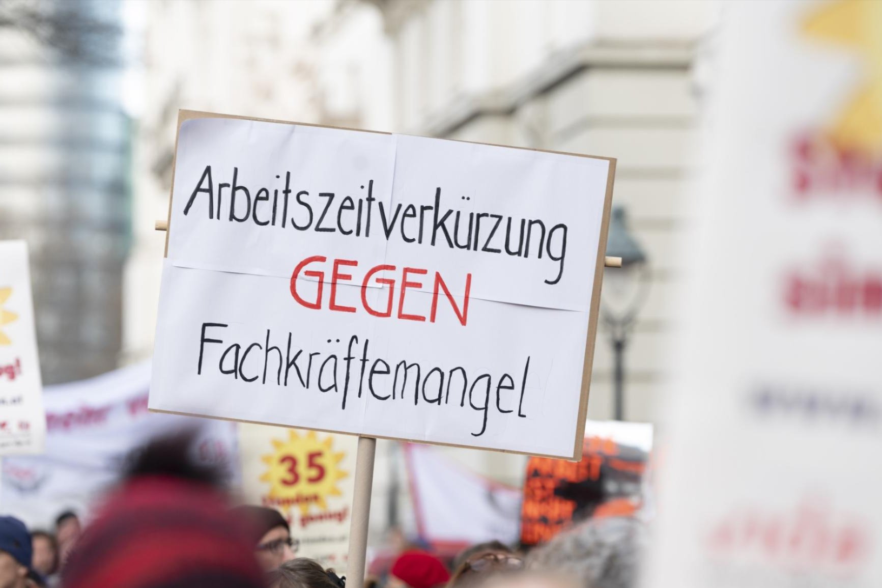 „Nahaufnahme eines Protest-Schildes auf einer Demonstration. Darauf steht: ‚Arbeitszeitverkürzung GEGEN Fachkräftemangel‘. Im Hintergrund weitere Demonstrierende mit Bannern.“