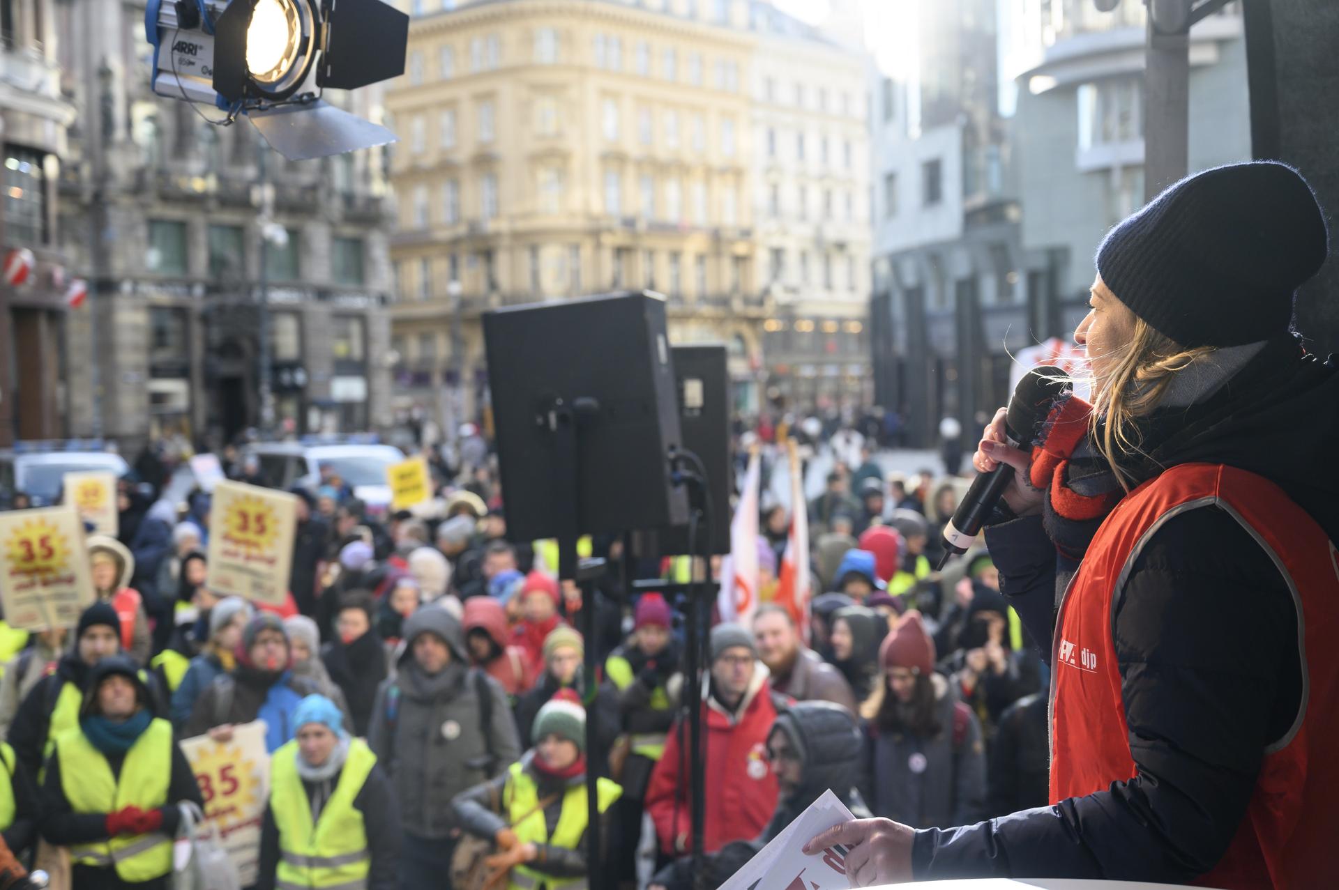 „Blick von der Bühne auf eine Demonstration in einer Innenstadt. Eine Frau im roten Überwurf spricht ins Mikrofon, vor ihr eine große Menschenmenge mit Transparenten und gelben Warnwesten. Auf den Schildern steht ‚35 Stunden sind genug‘.“